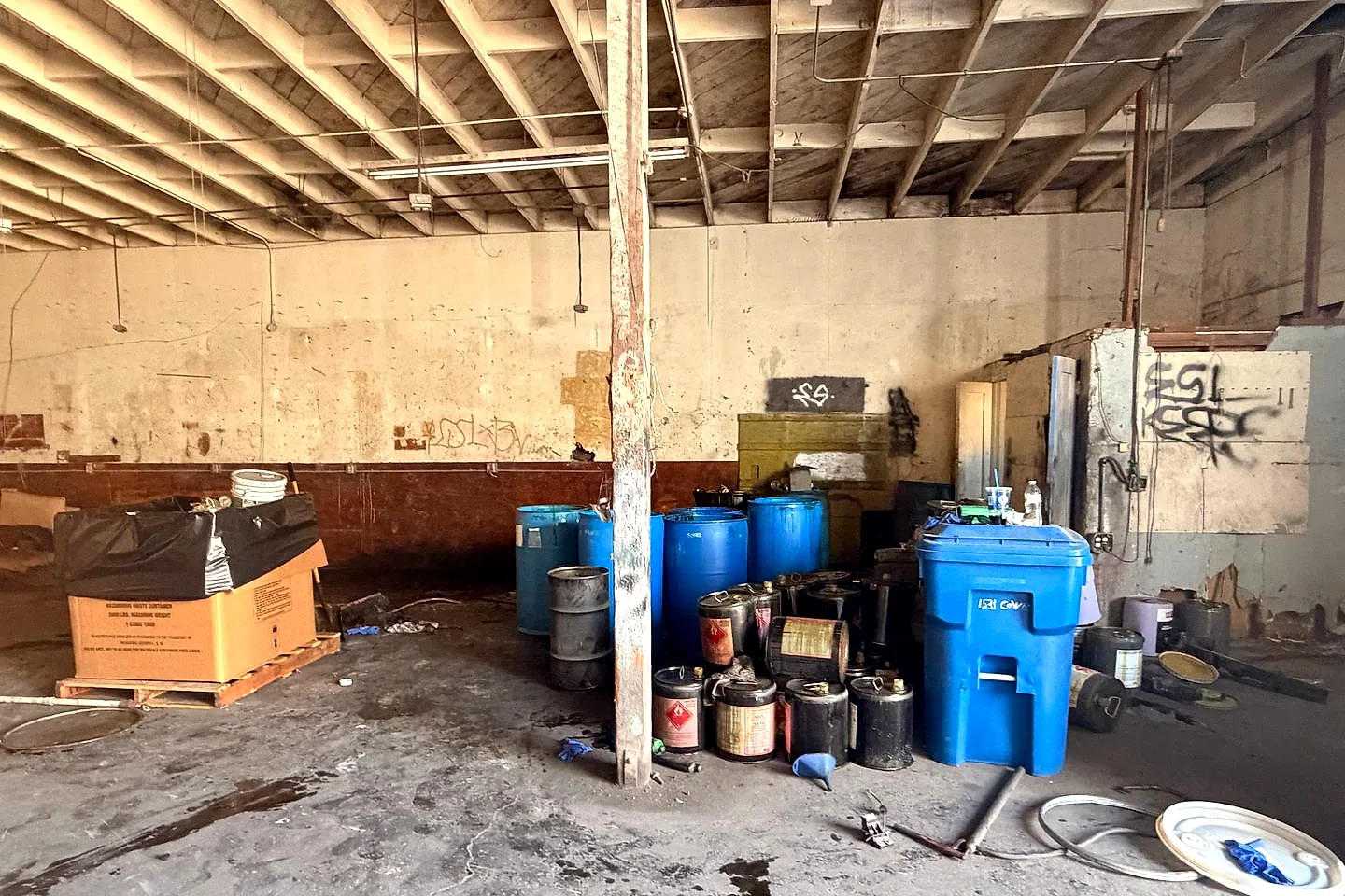 Vacant industrial building with blue drums, black steel drums, and a blue waste bin against the wall before cleanout
